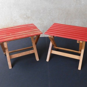 May include: Two red folding stools with wooden frames. The stools are made of wood and have a red painted finish.