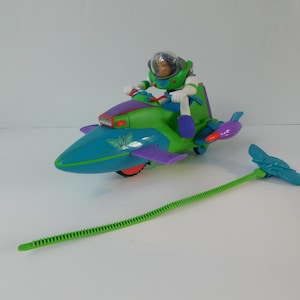 May include: A plastic action figure of a space ranger character with a clear helmet, seated on a green and blue rocket-shaped vehicle. The vehicle features purple wings, red accents, and a green ripcord mechanism with a blue handle attached to the side.