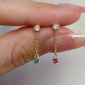 Könnte beinhalten: Ein Paar goldfarbene Hängeohrringe. Jeder Ohrring hat oben einen kleinen, runden, klaren Stein, der durch eine zarte Kette mit einem Edelstein unten verbunden ist. Ein Ohrring hat einen grünen Edelstein, der andere einen rosa.