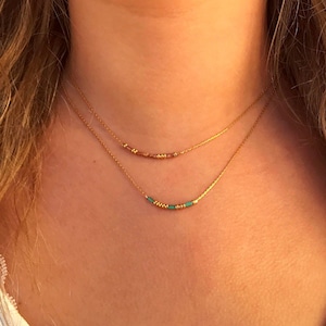 Puede incluir: Un conjunto de collares superpuestos que consta de dos delicadas cadenas doradas. La cadena superior presenta pequeñas cuentas marrones y doradas, mientras que la inferior muestra una secuencia de cuentas turquesas y doradas centradas.