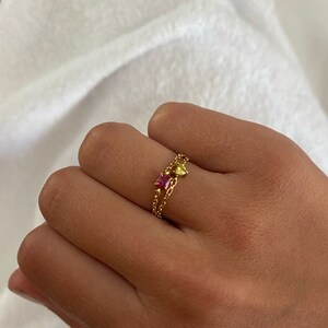May include: A gold ring with two linked bands, each with small beads. One band has a pink gemstone, the other a yellow gemstone. The ring is on a hand, against a white backdrop. The gemstones are likely small.