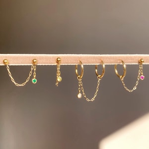 Puede incluir: Una colección de pendientes dorados exhibidos sobre una superficie de terciopelo rosa. Los pendientes incluyen estilos de tachuelas y aros, algunos con delicadas cadenas y colgantes de piedras preciosas de colores. Las piedras son verdes, amarillas, blancas y rosas.