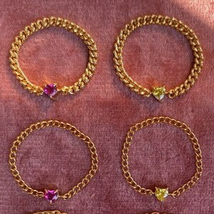 May include: Gold-colored chain rings with heart-shaped gemstones in pink, yellow, and blue. The rings are arranged on a dark pink surface, showcasing the jewelry's design and color variations. The rings are a simple yet elegant accessory.