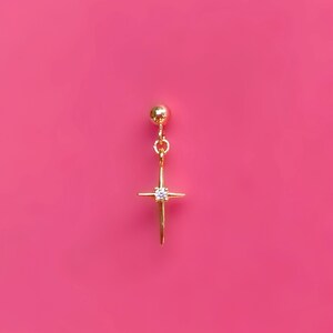 May include: A gold-colored earring with a starburst design and a small, clear gemstone at the center. The earring hangs from a small gold ball and is set against a bright pink background.