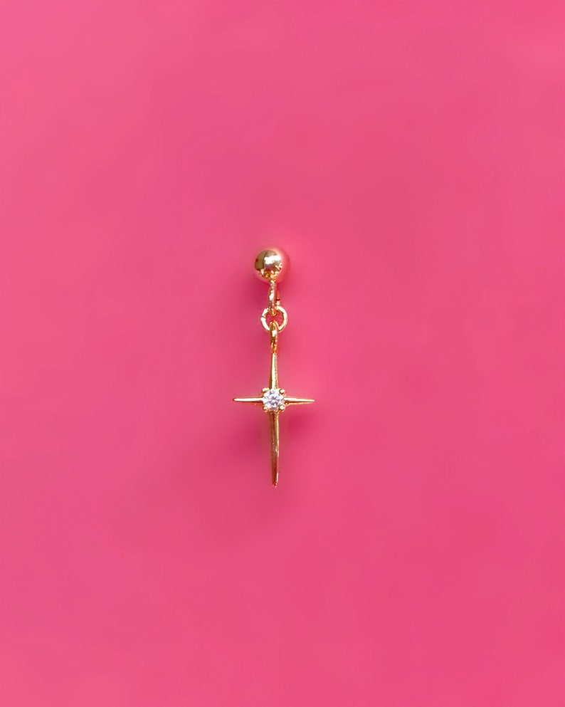 May include: A gold-colored earring with a starburst design and a small, clear gemstone at the center. The earring hangs from a small gold ball and is set against a bright pink background.