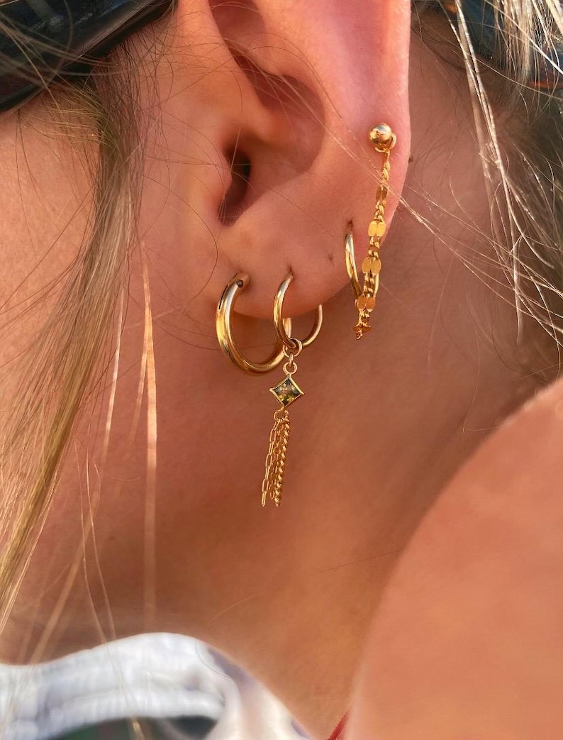 Op de afbeelding: Close-up van een oor versierd met meerdere gouden oorbellen. De oorbellen omvatten kleine hoepels, een bungelende oorbel met een vierkante groene edelsteen en ketting, en een delicate ketting van gouden schijven.