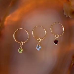 Op de afbeelding: Drie gouden oorringen, elk met een hartvormige edelsteen hanger. Eén oorring heeft een groene edelsteen, een andere een blauwe edelsteen en de derde een rode edelsteen. De oorbellen worden gepresenteerd op een bruin en beige oppervlak.