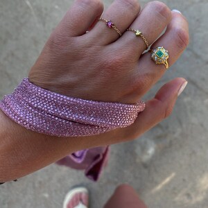 May include: A hand adorned with gold rings, including a chain ring with a pink gemstone and a ring with a green gemstone. A pink, sparkly bracelet wraps around the wrist. The image is taken outdoors.