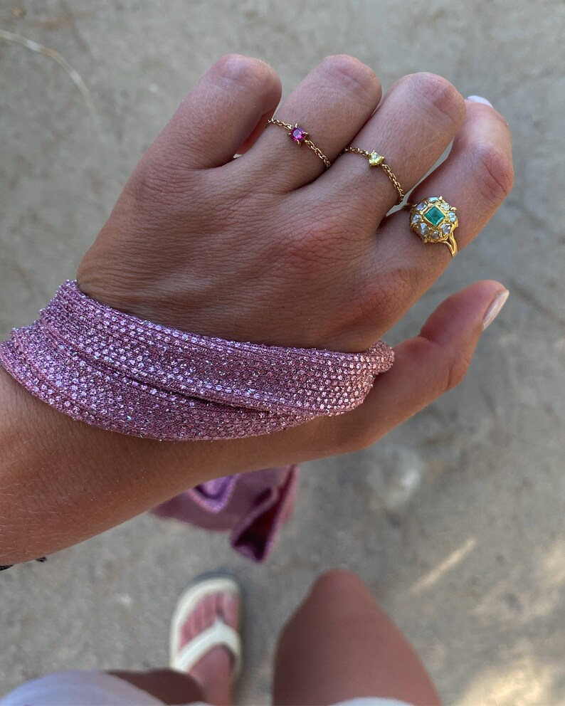 May include: A hand adorned with gold rings, including a chain ring with a pink gemstone and a ring with a green gemstone. A pink, sparkly bracelet wraps around the wrist. The image is taken outdoors.