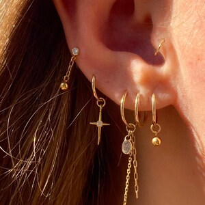 May include: A collection of gold earrings, including hoops, studs, and dangling styles. One earring features a cross pendant, while others have small charms and chain details. The earrings are displayed on an ear, showcasing their various designs.