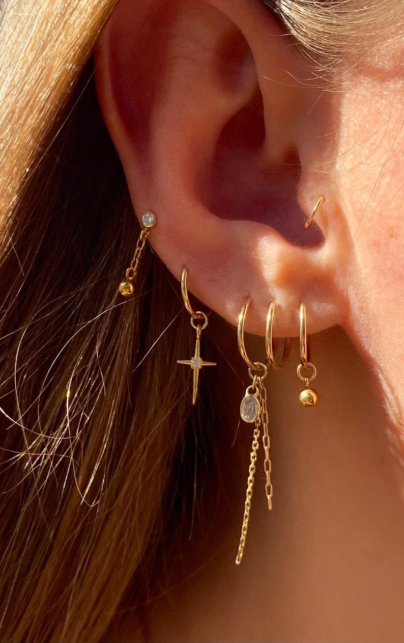 May include: A collection of gold earrings, including hoops, studs, and dangling styles. One earring features a cross pendant, while others have small charms and chain details. The earrings are displayed on an ear, showcasing their various designs.