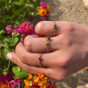 May include: Close-up of three gold chain rings with colorful gemstones. The rings feature a square emerald green stone, a pink square stone, and a small red stone. The rings are displayed against a backdrop of vibrant flowers.