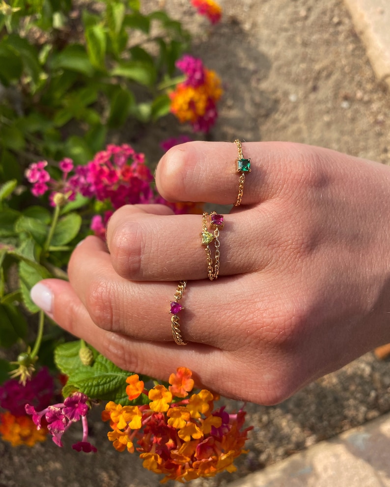 May include: Close-up of three gold chain rings with colorful gemstones. The rings feature a square emerald green stone, a pink square stone, and a small red stone. The rings are displayed against a backdrop of vibrant flowers.