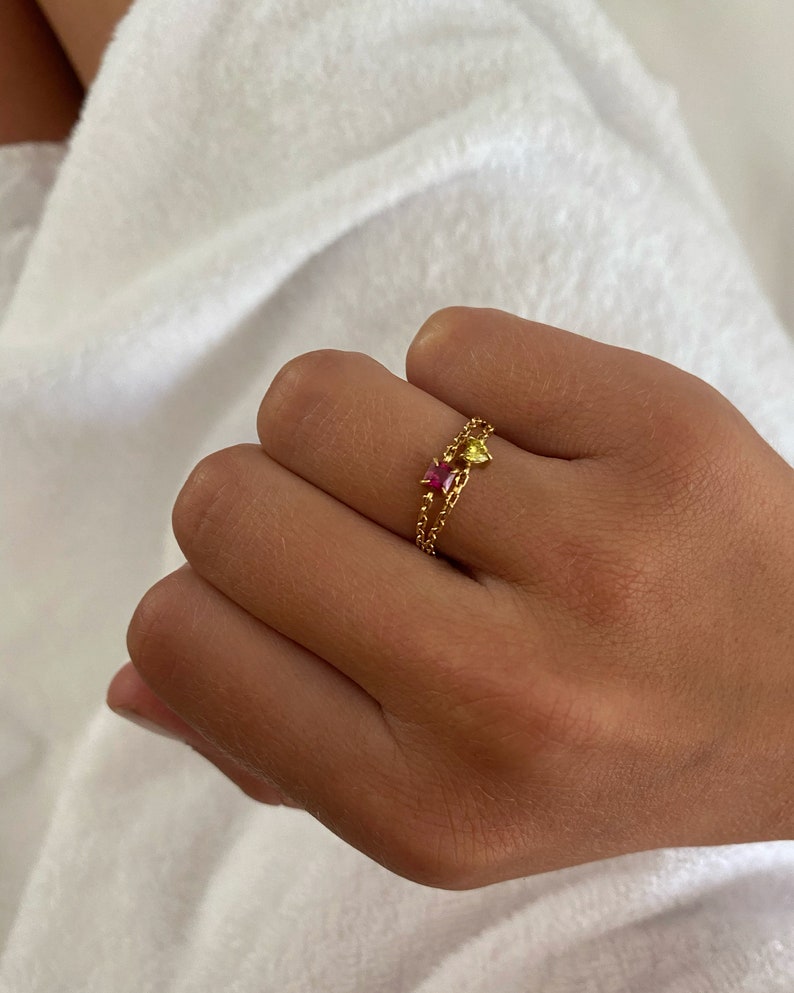 May include: A gold ring with two linked bands, each with small beads. One band has a pink gemstone, the other a yellow gemstone. The ring is on a hand, against a white backdrop. The gemstones are likely small.