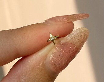 Puede incluir: Un pequeño pendiente dorado con un diseño de estrella de cuatro puntas. La estrella presenta una pequeña gema transparente en el centro. El pendiente se sostiene entre los dedos sobre un fondo suave de color melocotón.