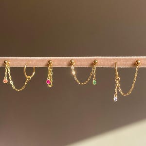 Op de afbeelding: Een verzameling goudkleurige oorbellen met ketting- en edelsteenaccenten. De oorbellen worden gepresenteerd op een roze fluwelen oppervlak. Sommige oorbellen hebben kleine, kleurrijke edelstenen, waaronder roze, groene en heldere stenen.