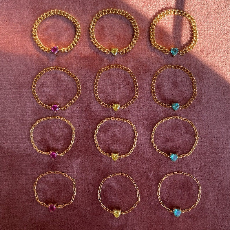 May include: Gold-colored chain rings with heart-shaped gemstones in pink, yellow, and blue. The rings are arranged on a dark pink surface, showcasing the jewelry's design and color variations. The rings are a simple yet elegant accessory.