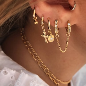 May include: An assortment of gold earrings, including small hoops, charm earrings with a cross and a pendant, and hoop earrings with a chain. The earrings are worn on the ear, with a gold necklace visible in the foreground.