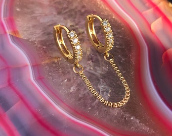 May include: Gold-toned double hoop earrings. Each hoop is adorned with small, clear gemstones. A delicate gold chain connects the two hoops, creating a stylish and modern look. The earrings are displayed against a pink and white background.