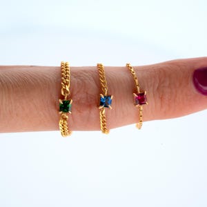 Op de afbeelding: Drie gouden kettingringen, elk met een vierkante edelsteen in groen, blauw en rood. De ringen worden op een vinger getoond tegen een witte achtergrond. De edelstenen zijn het middelpunt van de delicate sieraden.