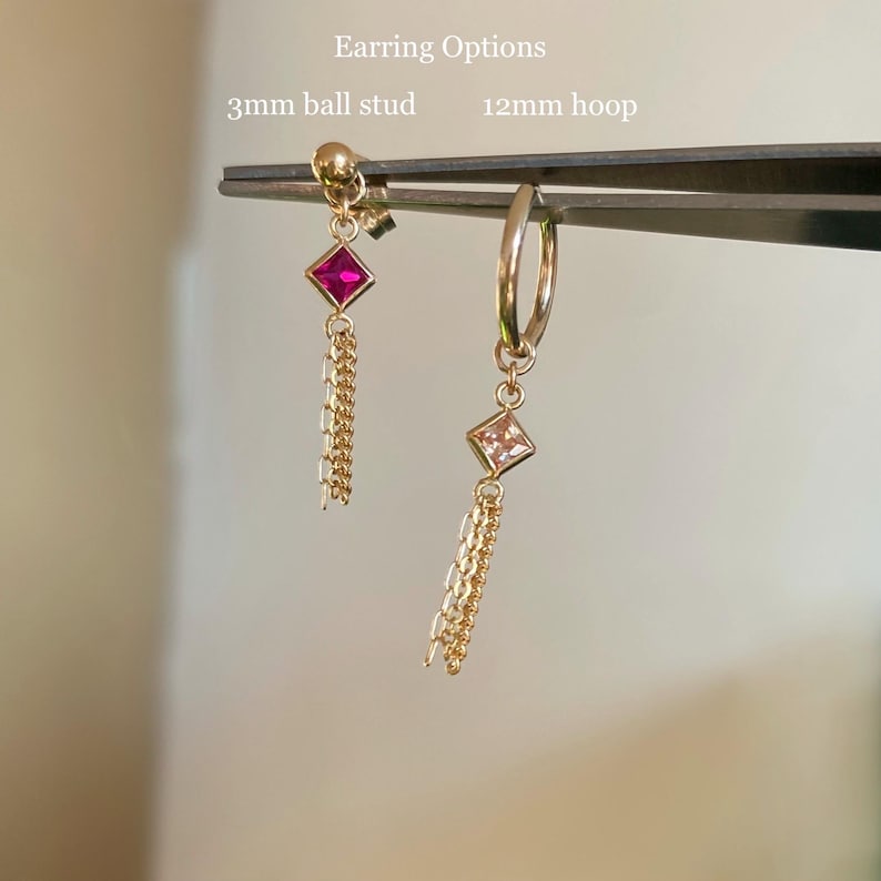 Może przedstawiać: Tekst Earring Options z kolczykiem sztyftem 3 mm z ciemnor&oacute;żowym kwadratowym kamieniem i złotym łańcuszkiem oraz kolczykiem kołem 12 mm z jasnor&oacute;żowym kwadratowym kamieniem i złotym łańcuszkiem, trzymanymi przez metalową pęsetę.