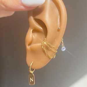 Op de afbeelding: Close-up van een oor versierd met meerdere gouden oorbellen. Een kleine oorring met een rechthoekige bedel met de letter "N" hangt aan de oorlel. Delicate kettingoorbellen en een oorstekers zijn ook zichtbaar.