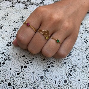 May include: A hand displays four gold rings. One ring features a pink gemstone, another has a heart pendant with the letter "H", and two others have green and yellow gemstones. The rings are on a white lace surface.