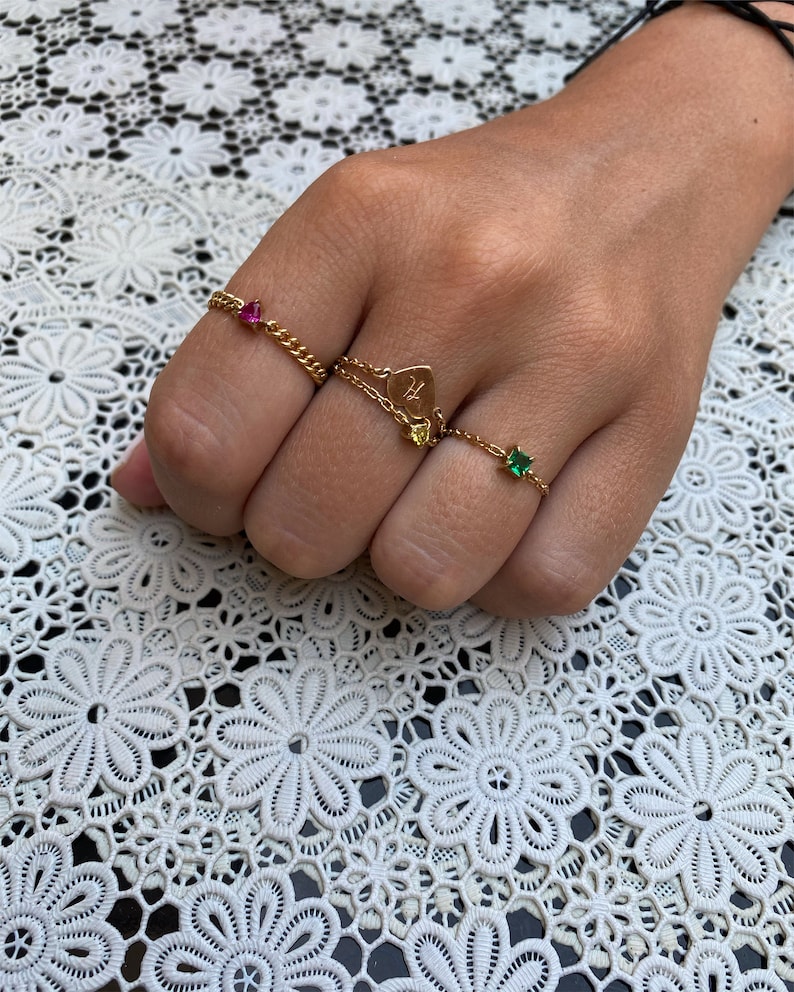 May include: A hand displays four gold rings. One ring features a pink gemstone, another has a heart pendant with the letter "H", and two others have green and yellow gemstones. The rings are on a white lace surface.