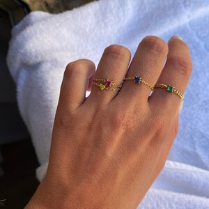May include: A hand wearing three gold chain rings, each with a different colored gemstone: yellow, red, blue, and green. The rings are set against a white background.