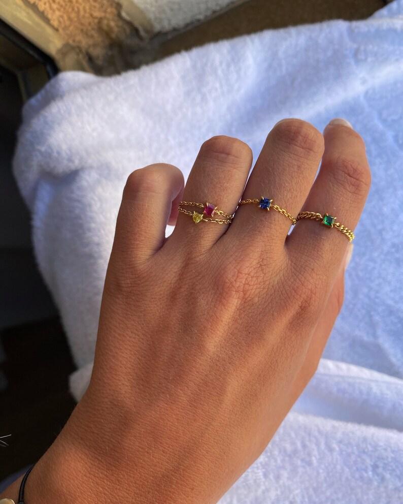 May include: A hand wearing three gold chain rings, each with a different colored gemstone: yellow, red, blue, and green. The rings are set against a white background.