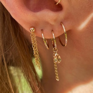 Puede incluir: Primer plano de una oreja adornada con múltiples pendientes dorados. La oreja presenta un pequeño pendiente de oro con una cadena, tres aros dorados de diferentes tamaños y un colgante en forma de lágrima colgando de una cadena.