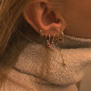 Peut inclure: Gros plan sur une oreille ornée de multiples boucles d'oreilles dorées. Les boucles d'oreilles comprennent de petits cerceaux, une chaîne pendante avec une pierre de couleur rubis et une chaîne délicate avec une petite perle dorée. L'oreille est posée sur un fond de pull gris.