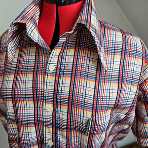 Puede incluir: Una camisa de manga corta con botones y un estampado de cuadros multicolores. La camisa tiene un cuello puntiagudo y un solo bolsillo en el pecho.