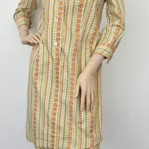 Op de afbeelding: Een vintage jurk op een mannequin. De jurk heeft een knoopsluiting, een kraag en lange mouwen. Het heeft een patroon van verticale strepen en cirkels in oranje, groen en wit. De jurk valt tot op de knieën, ongeveer 90 cm lang.