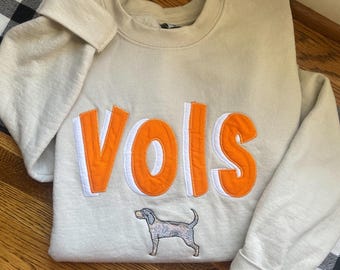 Op de afbeelding: Een licht beige sweatshirt met het woord "VOLS" geborduurd in oranje en wit. Onder het woord is een klein geborduurd beeld van een zwart-witte hond.