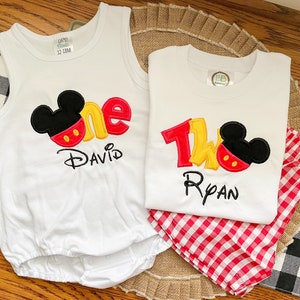 May include: Two white onesies with Mickey Mouse ears and the numbers one and two in red, yellow, and black. The onesies are personalized with the names "David" and "Ryan".