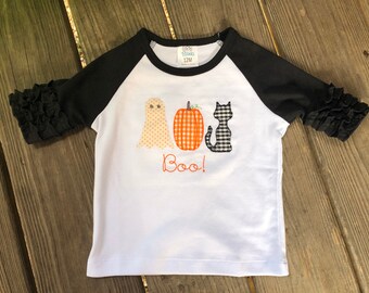 May include: White long-sleeve shirt with black raglan sleeves and a black and white gingham pumpkin, ghost, and cat applique. The shirt says "Boo!" in black embroidery.