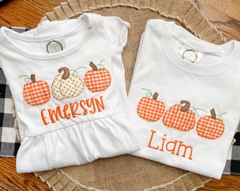 May include: Two white t-shirts with embroidered pumpkins and names. The shirt on the left has three pumpkins and the name "EMERSYN". The shirt on the right has three pumpkins and the name "Liam".