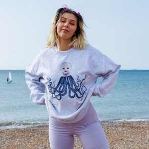 May include: A woman wearing a grey sweatshirt with a graphic of a baby's face inside an octopus. She is wearing lavender bike shorts and white sneakers. She is standing on a beach with the ocean in the background.