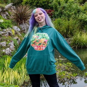 May include: A teal green hoodie featuring a colourful floral and fish design. The hoodie has a drawstring hood and a kangaroo pocket. The person wearing the hoodie has purple and blue hair.