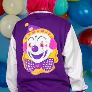 May include: A purple and white baseball jacket with a colorful clown face graphic on the back. The clown has a yellow hat, a pink bow, and a big smile.