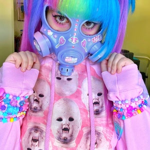 May include: A person with bright blue and pink hair wearing a pastel purple gas mask with stickers and a pink hoodie with a repeating pattern of doll faces.