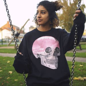 May include: A person wearing a black sweatshirt with a pink and white skull graphic. The skull has a cloudy sky inside of it. The person is sitting on a swing.