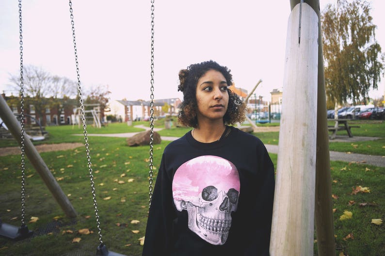 May include: A woman wearing a black sweatshirt with a pink and white skull graphic. The skull has a pink brain inside. She is standing in front of a swing set in a park.