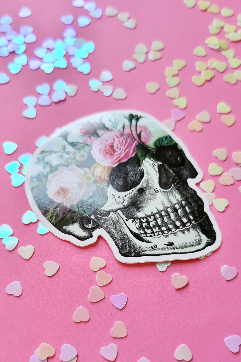 May include: A white skull sticker with a floral design. The skull is outlined in black and has a detailed anatomical drawing of the teeth and bones. The flowers are pink and white roses with green leaves. The sticker is on a pink background with iridescent heart-shaped confetti.