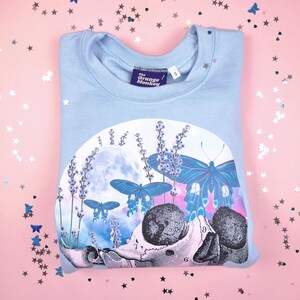 Pu&ograve; includere: Felpa blu chiaro con una stampa grafica di uno scheletro con farfalle e fiori di lavanda. Il testo "The Grunge Monkey" &egrave; stampato sull'etichetta.