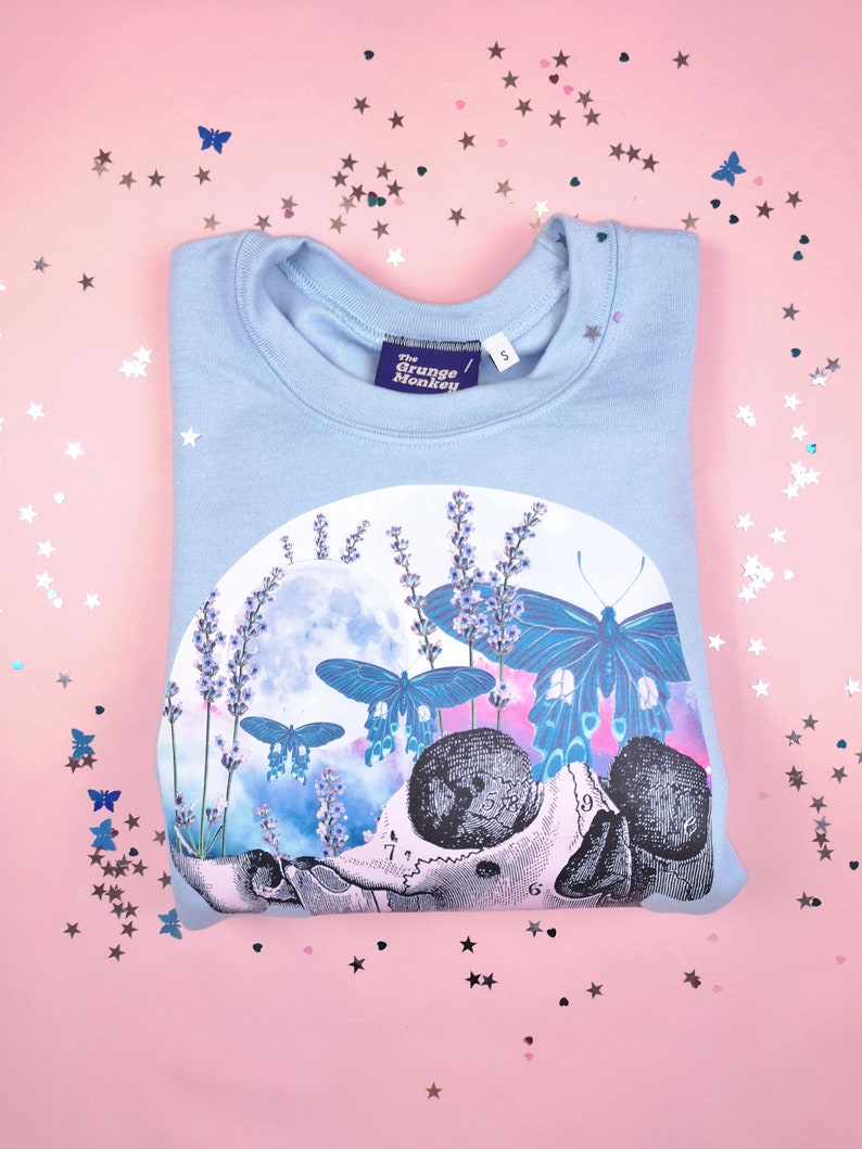 Pu&ograve; includere: Felpa blu chiaro con una stampa grafica di uno scheletro con farfalle e fiori di lavanda. Il testo "The Grunge Monkey" &egrave; stampato sull'etichetta.