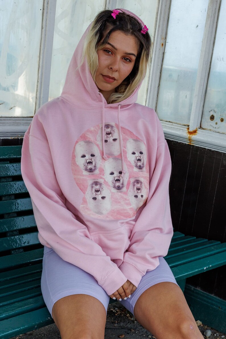May include: A pink hoodie with a graphic print of multiple baby faces on a pink background. The hoodie has a drawstring hood and a kangaroo pocket.