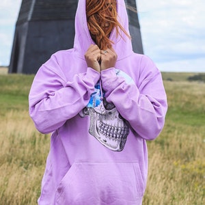 May include: A light purple hoodie with a skull graphic on the front. The hoodie has a drawstring hood and a kangaroo pocket. The person is wearing dark gray shorts. The background includes a windmill and a field of tall grass.