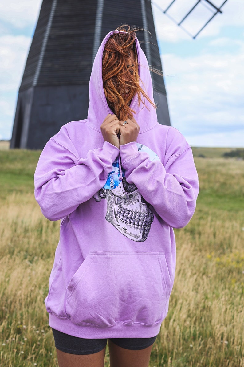May include: A light purple hoodie with a skull graphic on the front. The hoodie has a drawstring hood and a kangaroo pocket. The person is wearing dark gray shorts. The background includes a windmill and a field of tall grass.